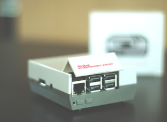 A small retro gaming console designed to resemble an old school entertainment system, featuring multiple USB ports on its front. The console is primarily white with some gray accents, and it sits on a dark surface. A blurred box with an image of the console is visible in the background.