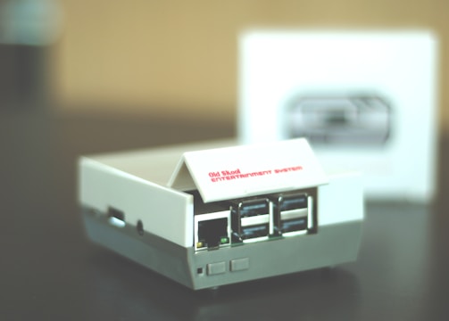 A small retro gaming console designed to resemble an old school entertainment system, featuring multiple USB ports on its front. The console is primarily white with some gray accents, and it sits on a dark surface. A blurred box with an image of the console is visible in the background.