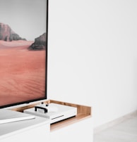 A modern interior setting featuring a white entertainment console with wooden accents. On the console, there is a gaming console with a controller on top. A large framed picture of a desert landscape with red sand and rocky formations is positioned next to the console.