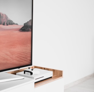 A modern interior setting featuring a white entertainment console with wooden accents. On the console, there is a gaming console with a controller on top. A large framed picture of a desert landscape with red sand and rocky formations is positioned next to the console.