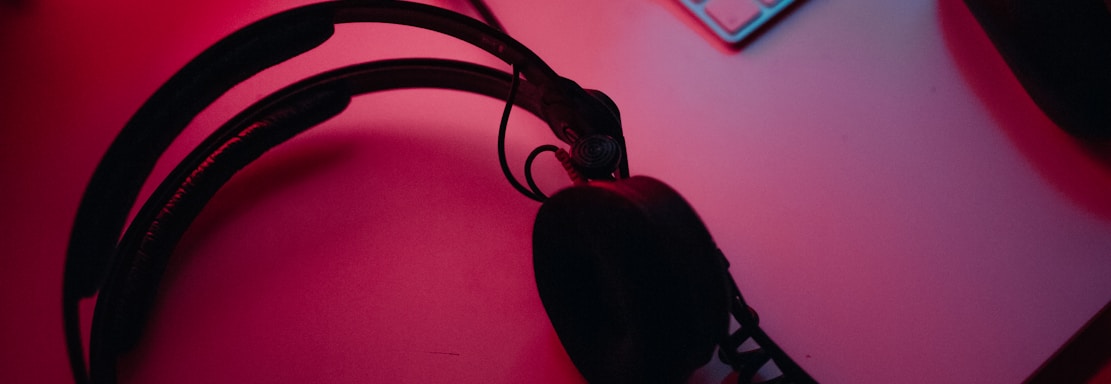 A dimly lit gaming setup with a monitor displaying a game's menu on the screen. A mechanical keyboard sits in front of the monitor. A pair of over-ear headphones and a game controller rest on the desk. The scene is bathed in a red and blue light, creating a moody gaming atmosphere.