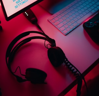 A dimly lit gaming setup with a monitor displaying a game's menu on the screen. A mechanical keyboard sits in front of the monitor. A pair of over-ear headphones and a game controller rest on the desk. The scene is bathed in a red and blue light, creating a moody gaming atmosphere.