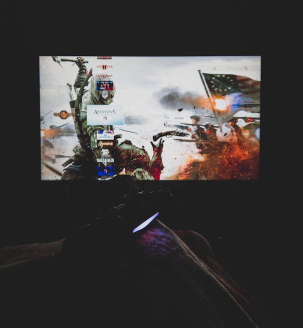 A dimly lit room with a television screen displaying a dynamic, action-packed video game interface featuring a historical battle scene. The foreground shows the silhouette of a gaming console controller held by a person.