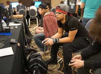 Several people are sitting in a room with gaming consoles and monitors, focusing intently on their screens. The setting appears to be a casual gaming event, with participants holding controllers while playing video games. The room has a casual atmosphere with chairs and tables set up for gaming.
