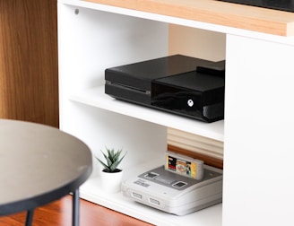 A black gaming console is situated on the middle shelf of a modern, white wooden entertainment unit. Below it, an older version of a gaming console sits with a game cartridge inserted. A small potted plant adds a touch of greenery next to the vintage console. The top of the unit is made of a light wood, and a flat-screen monitor is partly visible on it. The setup is minimalistic and clean, with a round black table in the foreground.