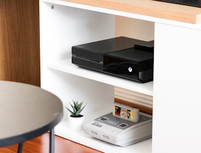 A black gaming console is situated on the middle shelf of a modern, white wooden entertainment unit. Below it, an older version of a gaming console sits with a game cartridge inserted. A small potted plant adds a touch of greenery next to the vintage console. The top of the unit is made of a light wood, and a flat-screen monitor is partly visible on it. The setup is minimalistic and clean, with a round black table in the foreground.