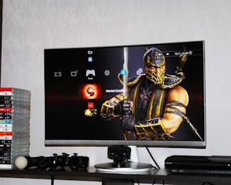 A flat-screen TV displays a video game interface featuring a character from Mortal Kombat in the center. Beside the TV is a stack of video game cases for the PlayStation 3, including titles like FIFA 14 and Grand Theft Auto. Below the TV, there is a gaming console, controllers, and other gaming equipment.