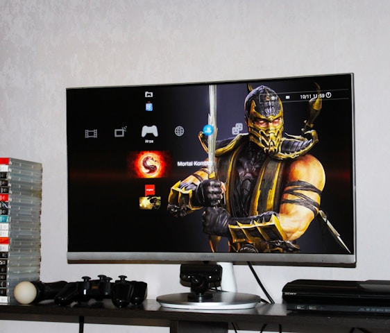 A flat-screen TV displays a video game interface featuring a character from Mortal Kombat in the center. Beside the TV is a stack of video game cases for the PlayStation 3, including titles like FIFA 14 and Grand Theft Auto. Below the TV, there is a gaming console, controllers, and other gaming equipment.