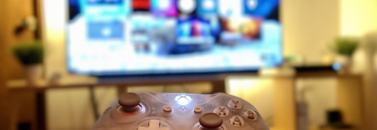 A gaming controller held in the foreground with a blurred television displaying a home screen interface in the background. The room has a cozy atmosphere with plants and a wooden shelf.