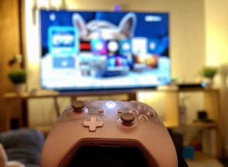A gaming controller held in the foreground with a blurred television displaying a home screen interface in the background. The room has a cozy atmosphere with plants and a wooden shelf.
