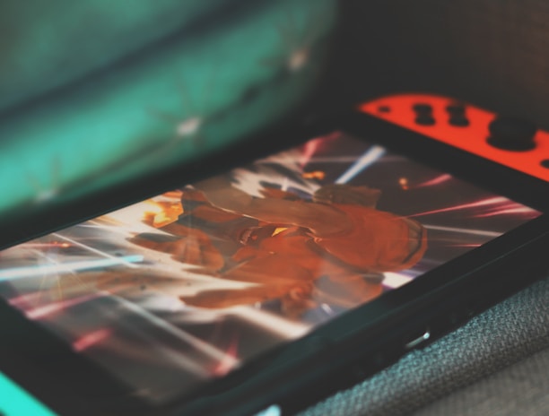 A portable gaming console with neon red and blue controllers displays an action-packed scene from a video game. The screen is focused on a character surrounded by bright, dynamic light effects.