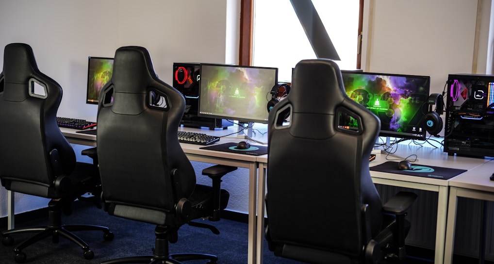 A row of gaming stations equipped with large monitors, gaming chairs, keyboards, and headphones. The setup is organized and ready for gaming, featuring a colorful screen display. The room has a modern and tech-savvy ambiance.