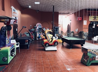 An indoor arcade area featuring various gaming machines, including an air hockey table and a ride-on toy car. The room has a tiled floor and several colorful gaming devices scattered around. Chairs and arcade signage are visible, creating a playful and entertainment-focused atmosphere.