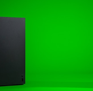 A black, rectangular gaming console stands upright against a vibrant green background. The console has a simple, sleek design with minimalistic features including a disc slot and a few buttons.