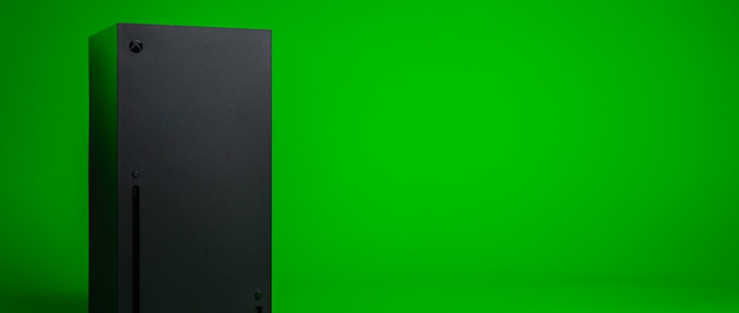 A black, rectangular gaming console stands upright against a vibrant green background. The console has a simple, sleek design with minimalistic features including a disc slot and a few buttons.