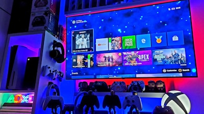 A modern gaming setup featuring a large wall-mounted screen displaying a gaming console interface. The room is decorated with gaming-themed items such as controllers and collectibles. The ambient lighting includes shades of blue and red, creating a vibrant atmosphere.