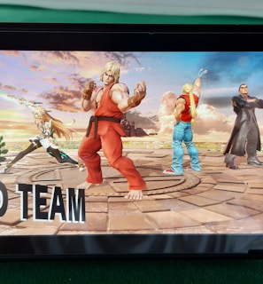 A handheld gaming console displays a screenshot of a fighting game featuring cartoonish characters in dynamic poses. The characters are standing in a virtual arena setting with a sky and cloud backdrop. The console is placed on a green surface.