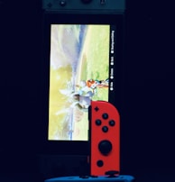 A gaming console with a screen displaying a video game scene, accompanied by two controllers, one colored red and the other blue. The devices are set against a dark backdrop, emphasizing the vibrant colors of the controllers.