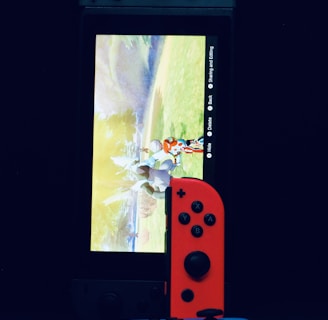 A gaming console with a screen displaying a video game scene, accompanied by two controllers, one colored red and the other blue. The devices are set against a dark backdrop, emphasizing the vibrant colors of the controllers.
