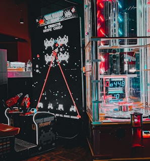 An arcade setting featuring a classic Space Invaders game setup and a modern gaming machine with seats. Bright neon lights illuminate the area, with scores displayed on digital screens. A bright and visually engaging environment full of electronic components.