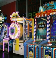 A row of brightly colored arcade game machines is arranged in a line against the wall. Each machine features vibrant lights and designs, with themes ranging from claw games to game show-inspired setups. The area has a playful and inviting atmosphere, suitable for entertainment or amusement centers.