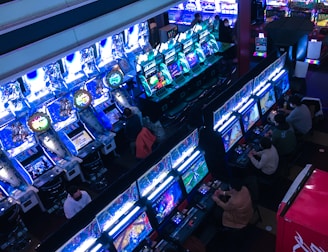 A bustling arcade filled with rows of gaming machines, each with vibrant displays and controls. Several people are seated at these machines, deeply engaged in playing games. Bright fluorescent lights cast a blue and neon glow across the room, enhancing the lively and energetic atmosphere.