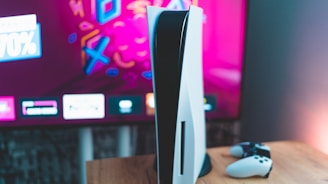 A gaming console is placed on a wooden table with a game controller nearby. In the background, a television screen displays colorful graphics, including geometric shapes and vibrant colors.