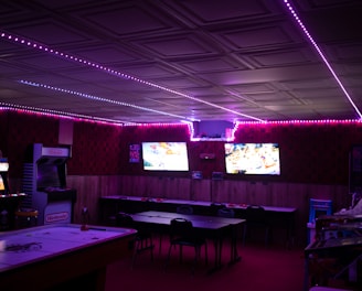 A dimly lit room with vibrant purple LED strip lights on the ceiling, featuring multiple gaming stations. There are two large screens showing video games, along with various arcade machines, including a Nintendo arcade cabinet. An air hockey table is positioned in the foreground.
