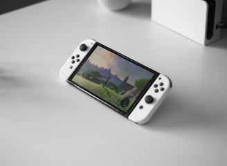 A portable gaming console with a screen showing a video game landscape, resting on a light-colored surface. The console has white controllers attached to either side of the screen. Nearby, there seems to be a white docking station or accessory.