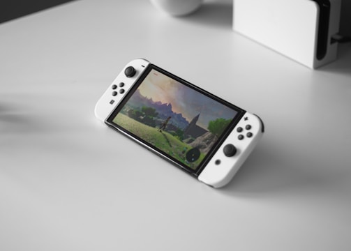 A portable gaming console with a screen showing a video game landscape, resting on a light-colored surface. The console has white controllers attached to either side of the screen. Nearby, there seems to be a white docking station or accessory.
