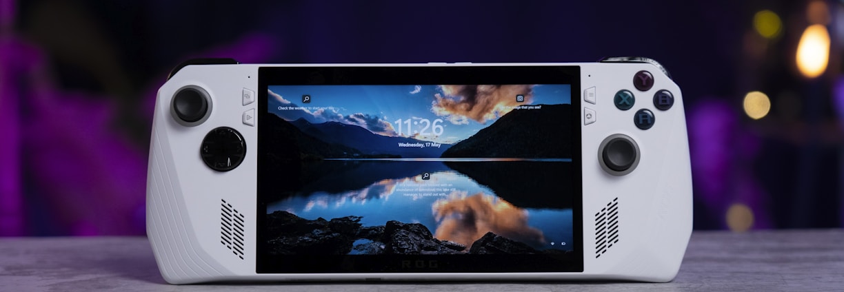 A handheld gaming console with a central screen displaying a scenic wallpaper of mountains and a lake, flanked by controller buttons and joysticks on both sides. The lighting around is dim, with purplish hues in the background, creating a modern and elegant atmosphere.
