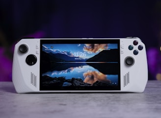 A handheld gaming console with a central screen displaying a scenic wallpaper of mountains and a lake, flanked by controller buttons and joysticks on both sides. The lighting around is dim, with purplish hues in the background, creating a modern and elegant atmosphere.