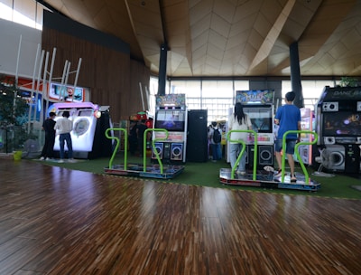 A vibrant arcade setting with several gaming machines lined up, including dance and rhythm game setups. People are engaged with the games, absorbed in their activities. The flooring is wooden, and the ceiling has a geometric design. Natural light filters through large windows, creating a lively atmosphere.