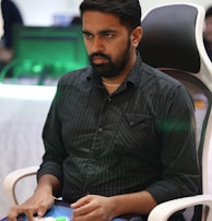A person is seated in a chair, focused intently on something, with hands positioned on a gaming console or arcade controller. The setup appears to be indoors, with visible screens and lighting in the background, suggesting an electronic gaming environment.